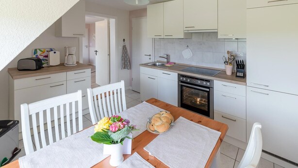 Una cocina compacta con armarios blancos, horno empotrado y una zona de comedor con una mesa para dos.