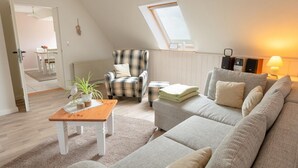 Un loft acogedor con techo alto, techo inclinado con tragaluz, un sillón escocés, un sofá gris, una mesa de centro de madera y un área de comedor al fondo.