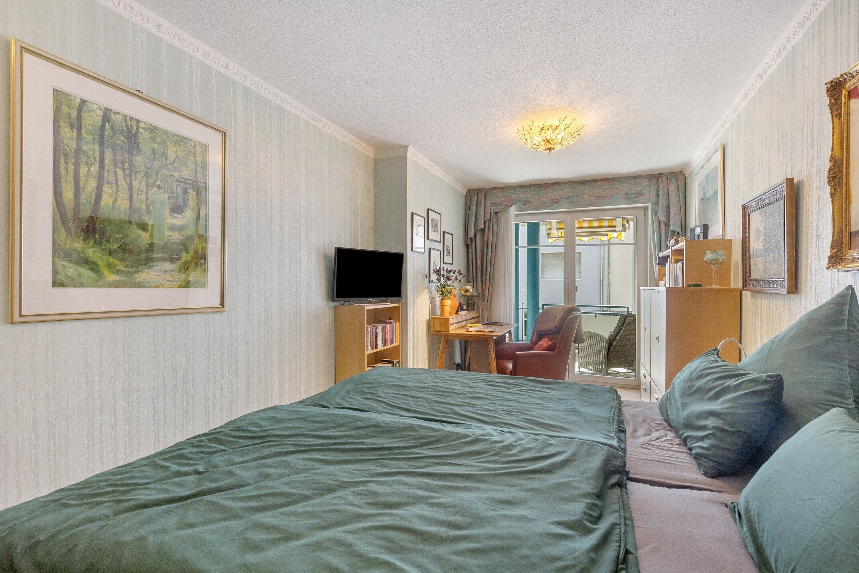 A hotel room with a bed, a desk with a chair, a television, and framed artwork on the walls.