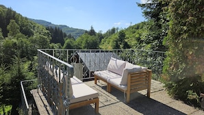 Un balcon doté d’un canapé et d’une table, entouré d’une rambarde en métal, et offrant une vue sur une zone boisée.