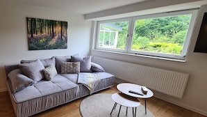 Un salon moderne avec un canapé d’angle, une petite table ronde, une grande fenêtre donnant sur un paysage verdoyant et un tableau accroché au mur.