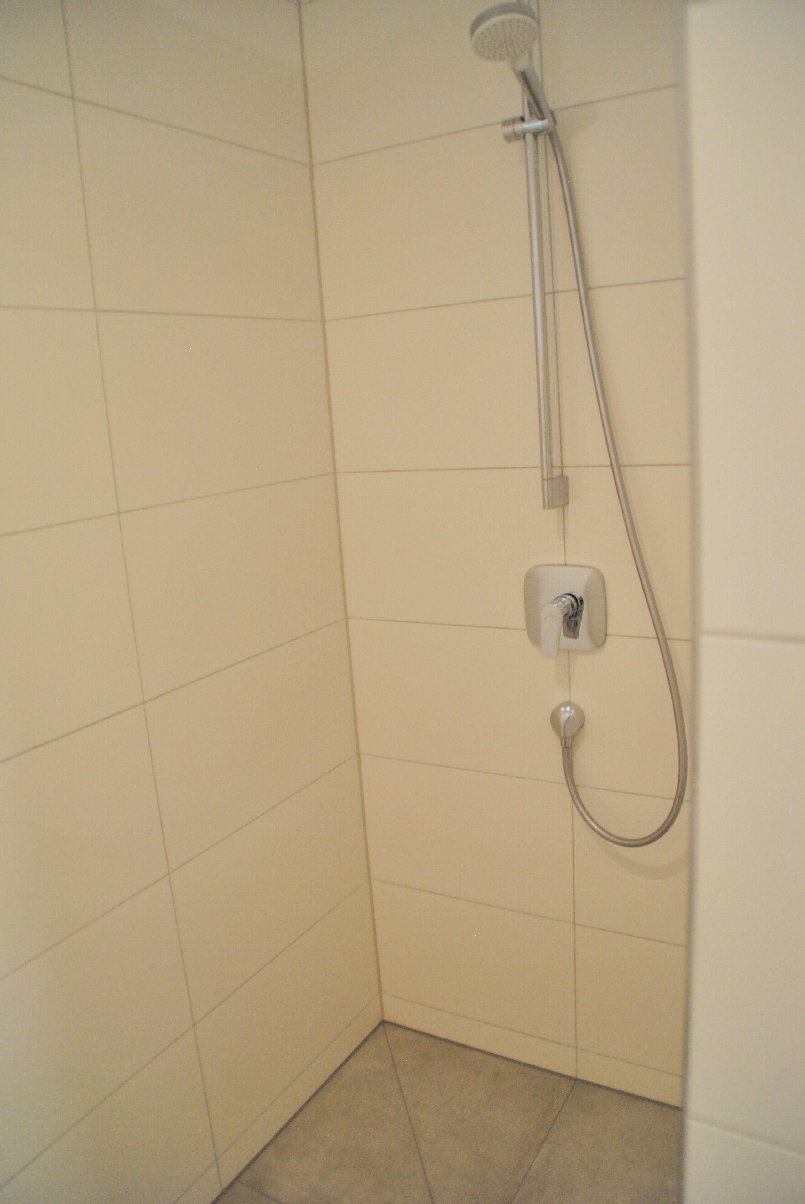 Un angolo di un bagno con pareti e pavimento piastrellati di bianco, un soffione e una manopola di regolazione.