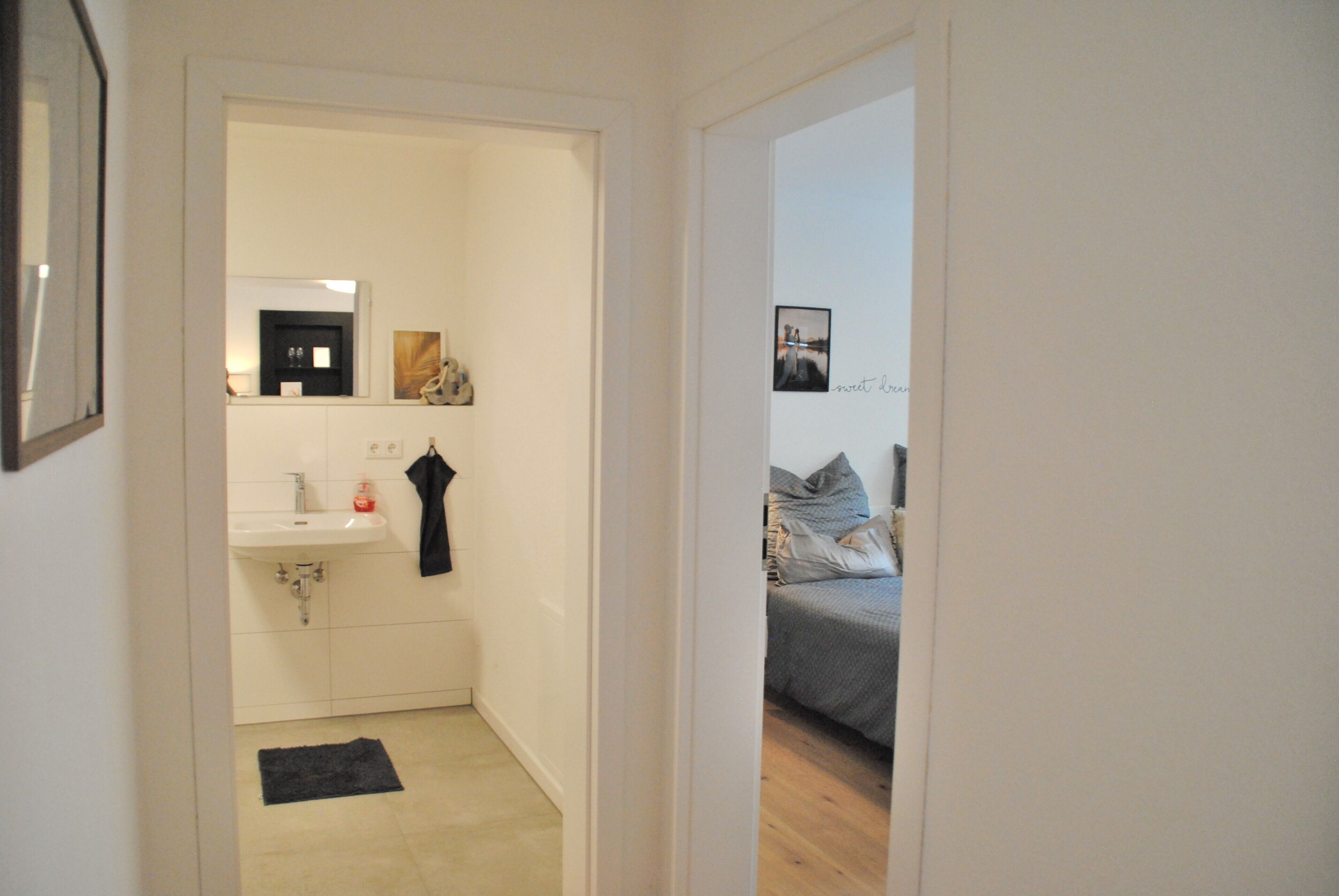 A narrow hallway leading to a bathroom with a sink and mirror, and a bedroom with a bed and pillows.