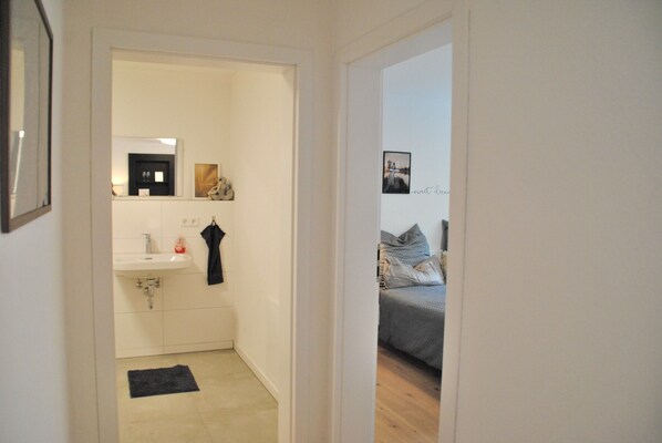 A narrow hallway leading to a bathroom with a sink and mirror, and a bedroom with a bed and pillows.