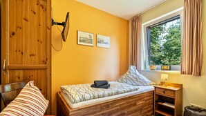 A room with a wooden bed, a bedside table with a lamp, a window with curtains, and two framed pictures on the wall.
