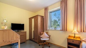 Ein Zimmer mit Fernseher, Holzschrank, Stuhl, Schreibtisch und einem Fenster mit Vorhängen.