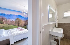 Hotellihuone, jossa on sänky, kylpyhuone, jossa on pesuallas ja peili, sekä kaunis näköala vuorille ja metsälle ikkunasta.