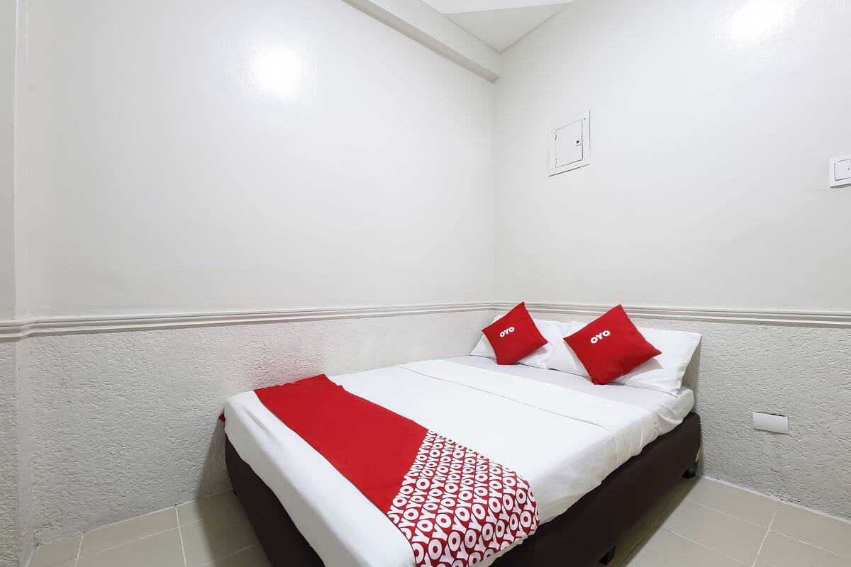 Una cama bien hecha con ropa de cama blanca y roja, dos almohadas rojas y un plaid estampado.