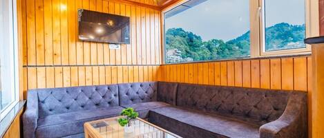 A cozy room with a wooden ceiling, a large window with a view of green hills, a grey sofa, and a glass coffee table.