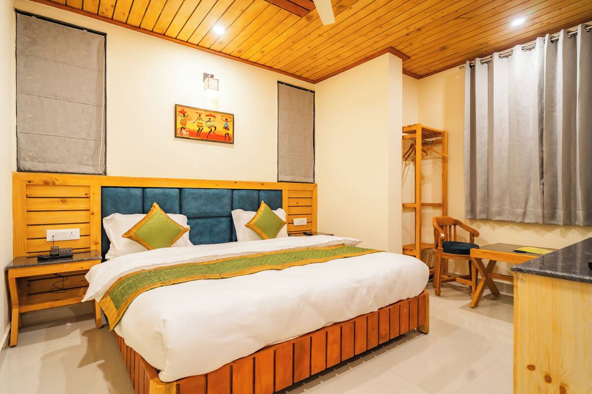 Un dormitorio con cabecera de madera, cama con ropa de cama blanca y almohadas verdes, mesita de noche, una silla de madera y un escritorio con silla.