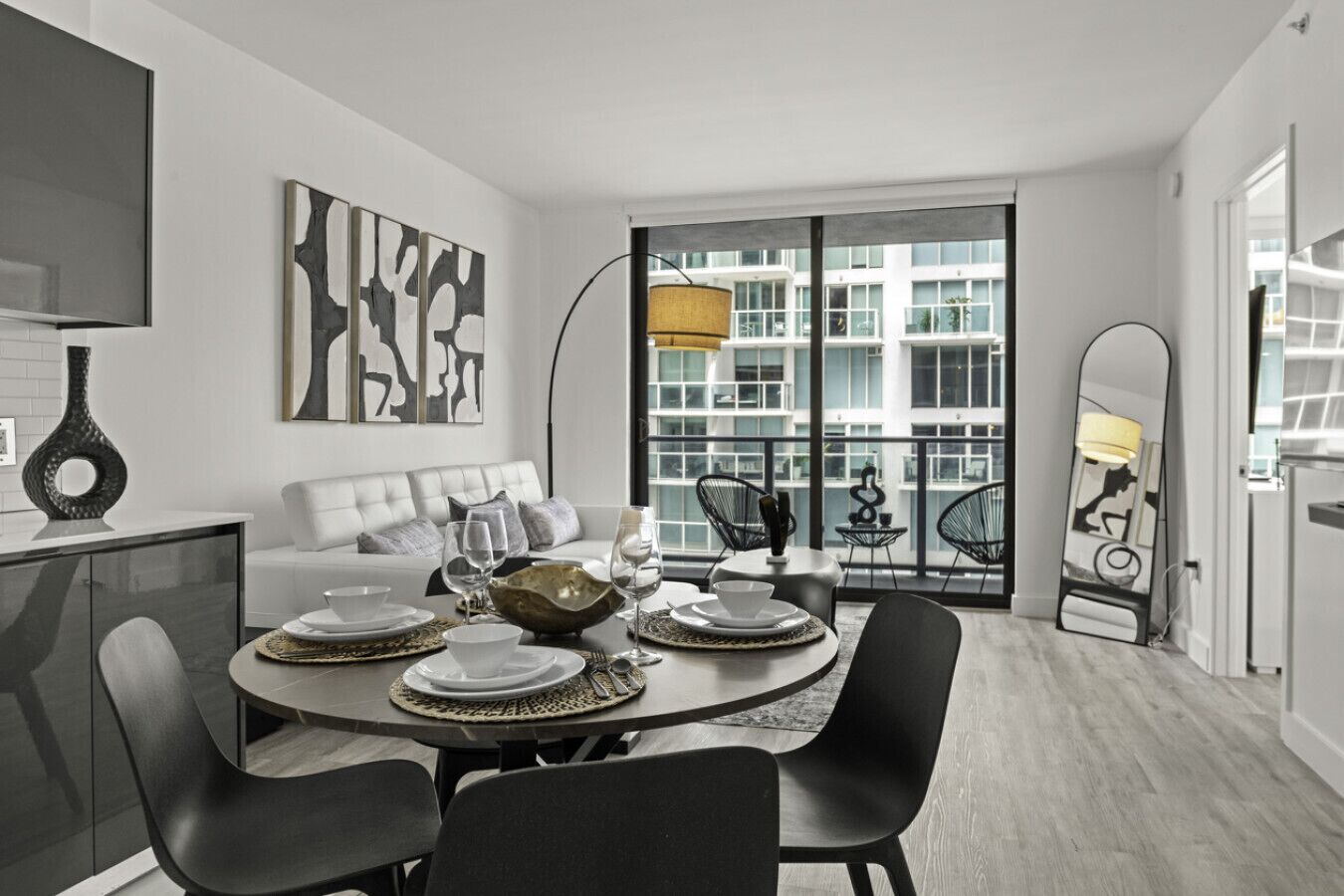 Un comedor moderno con una mesa redonda dispuesta para cuatro, un sof á y un amplio ventanal con vistas a edificios.