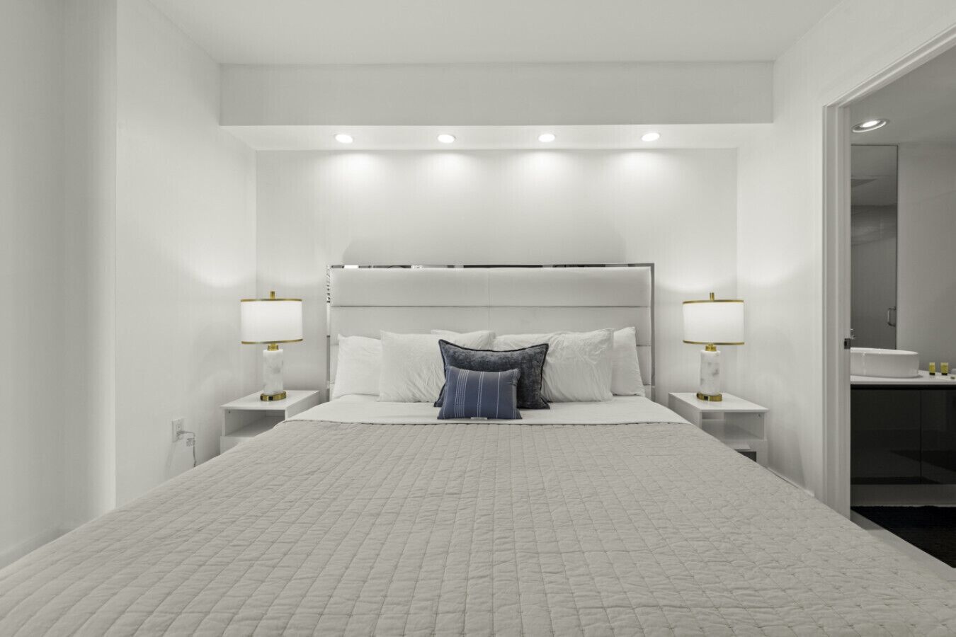 A modern hotel bedroom with a large bed, two bedside tables with lamps, and a view into the bathroom.