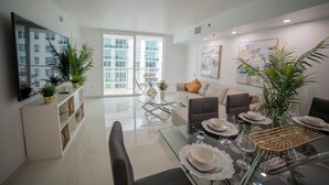 A modern living room with a glass dining table set for six, a sofa, and a TV mounted on the wall. - Cozy 4BR Apt@Brickell, free parking (Miami)