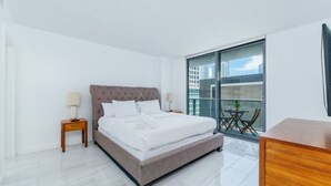 A modern bedroom with a large bed, a nightstand, a wooden dresser, and a balcony with a glass door. - Cozy 4BR Apt@Brickell, free parking (Miami)