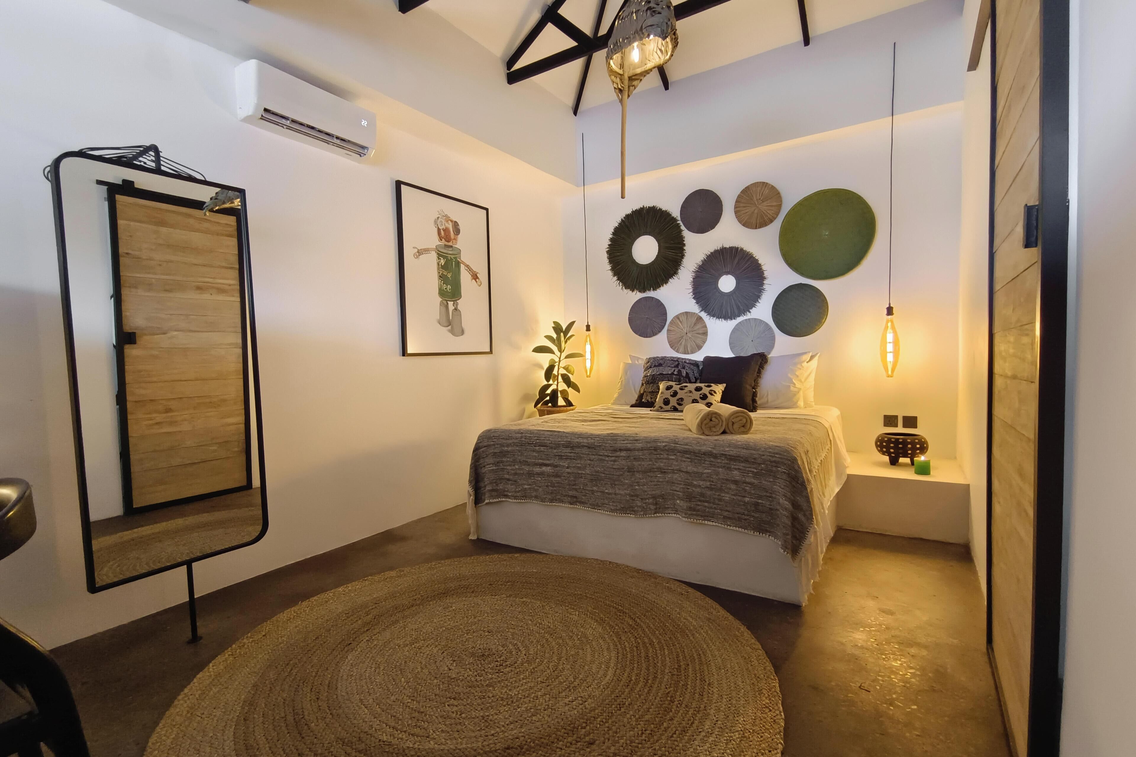 A bedroom with a bed, a wall-mounted mirror, a framed picture, decorative plates, a bedside lamp, and a small table with a plant.