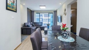 A modern living room with a sofa, a dining table with chairs, and a large window with a view of buildings. - Studio Apartment by Away Homes Rentals (Syokimau)