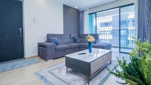 A modern living room with a grey sectional sofa, a glass coffee table, and a large window offering a view of buildings. - Studio Apartment by Away Homes Rentals (Syokimau)
