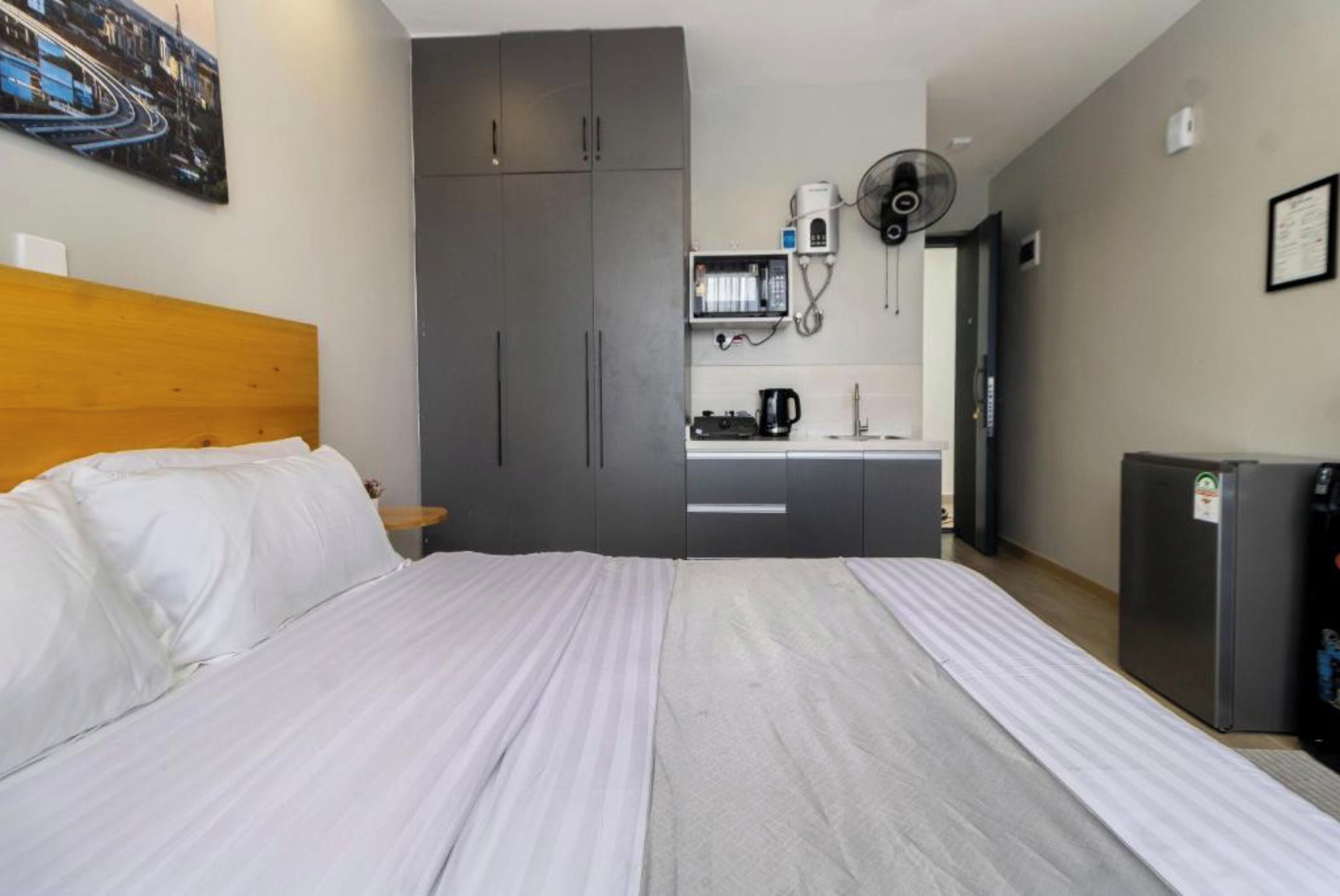 A hotel room with a bed, a kitchenette, a grey wardrobe, a small fridge, and a wall-mounted fan.