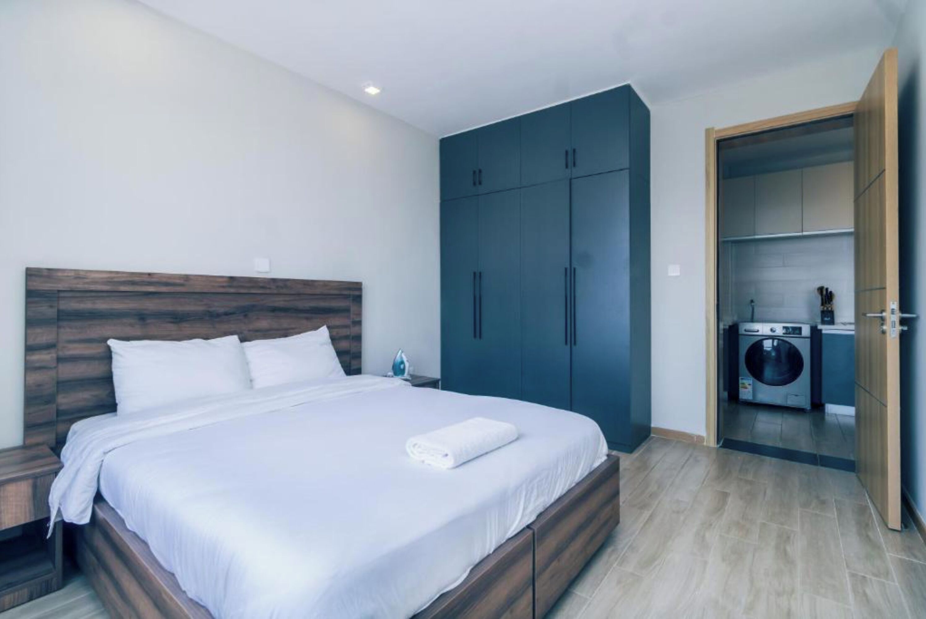 A bedroom with a wooden headboard, a large bed with white bedding, a dark blue wardrobe, and a laundry area with a washing machine.