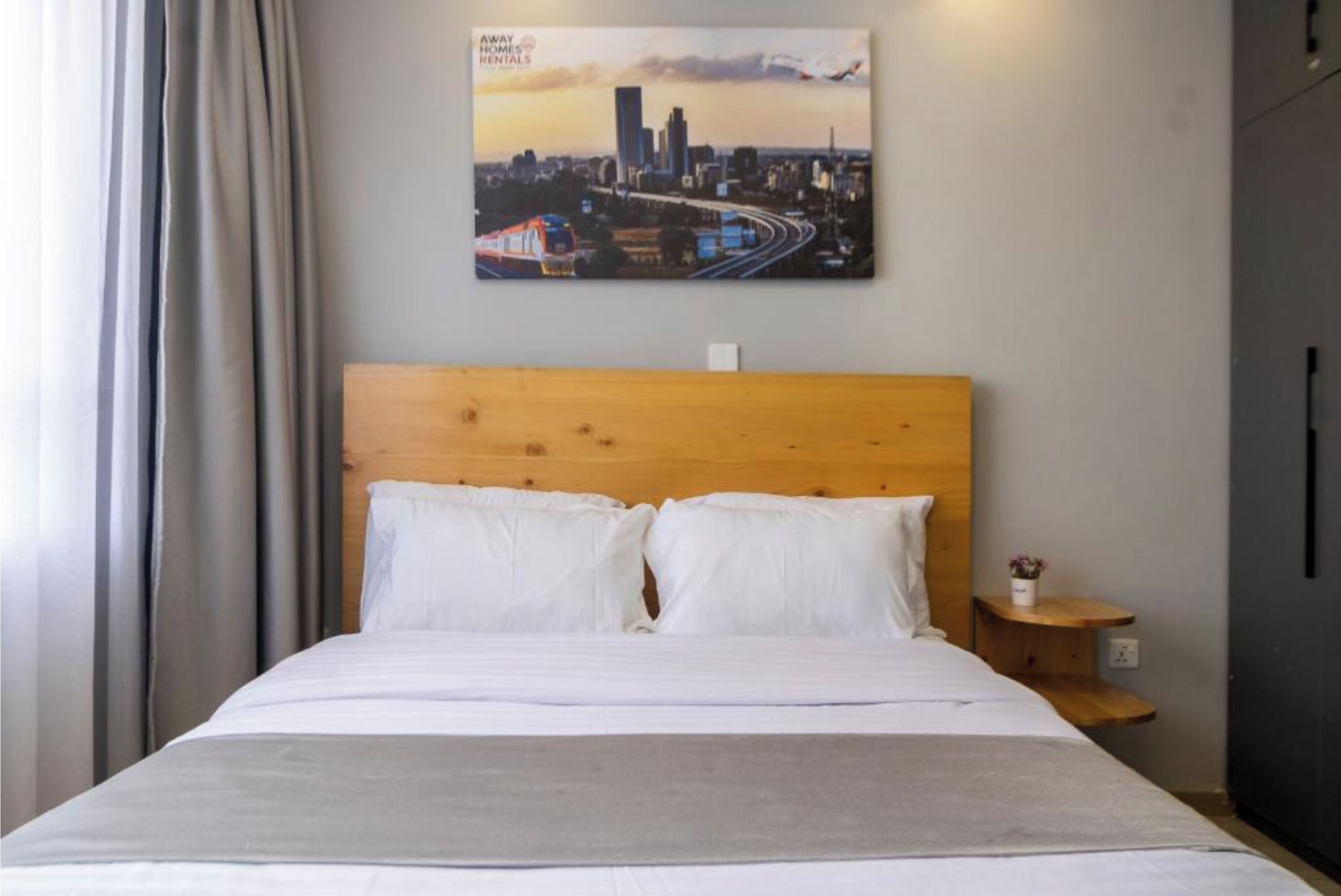A hotel room with a wooden headboard, a bed with white linens, a nightstand with a small vase, and a cityscape painting on the wall.