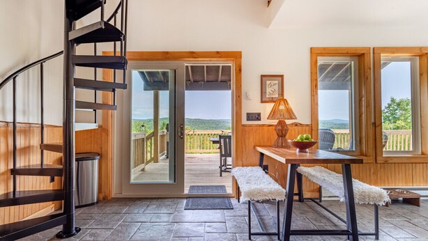 A spacious room with a spiral staircase, a wooden bench, a table with a lamp, and a view of the outdoors through a glass door. - Tiny Home with Spectacular Views & FP (Parksville)