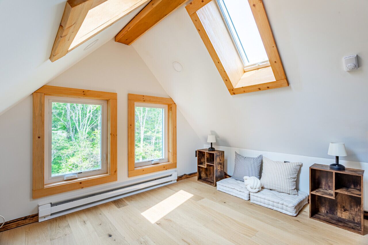 A cozy attic bedroom with a sloped ceiling, two large windows, a white sofa, and a wooden nightstand.