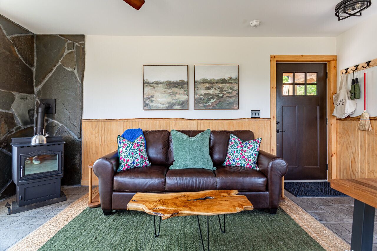 A cozy living room with a stone wall, a fireplace, a brown leather sofa with colorful cushions, and two landscape paintings on the wall.