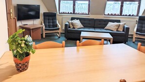 Un salon chaleureux avec un canapé foncé, deux fauteuils, une table basse en bois et un téléviseur posé sur un meuble. Il y a deux fenêtres avec des rideaux, un luminaire au plafond et une plante en pot sur une table.