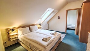 Une chambre avec un plafond en pente, un lit recouvert de draps blancs, une lampe de chevet et une vue sur une autre pièce, visible à travers une porte.