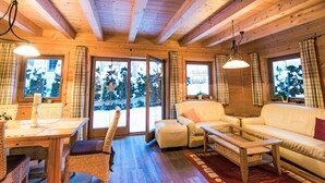 Acogedor interior de cabaña de madera con comedor, dos sofás y ventanas grandes con cortinas.