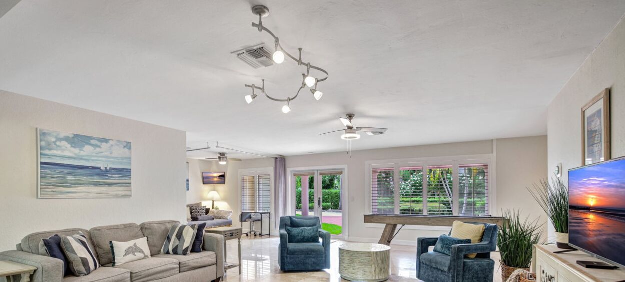A spacious living room with a large sofa, two armchairs, a coffee table, and a TV. There is a ceiling fan, a chandelier, and a painting on the wall.
