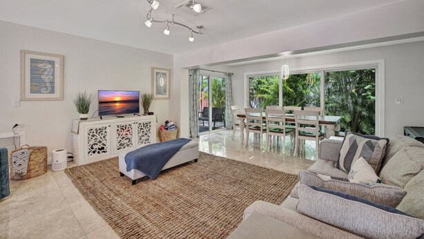 A spacious living room with a large TV, a dining area with a table and chairs, and a view of greenery through large windows. - Stylish 2br/3ba w-Heated Pool + Beach Walkable (Lauderdale-by-the-Sea)
