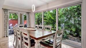 Un espace repas avec une table en bois et des chaises blanches, de grandes fenêtres donnant sur un paysage verdoyant et un luminaire suspendu.