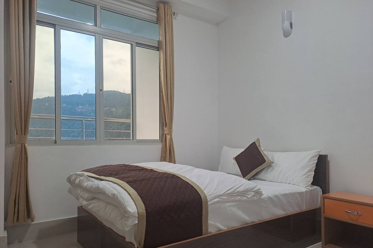 A bedroom with a bed, a nightstand, a window with curtains, and a view of a distant landscape.