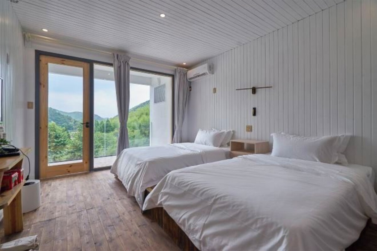 A bedroom with two beds, wooden flooring, a large window with a view of greenery, and a ceiling with recessed lighting.