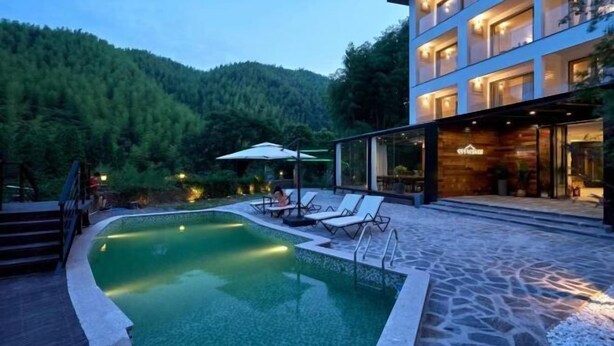 A modern hotel with a swimming pool, outdoor seating, and a forested backdrop. - Rest Valley (Deqing)