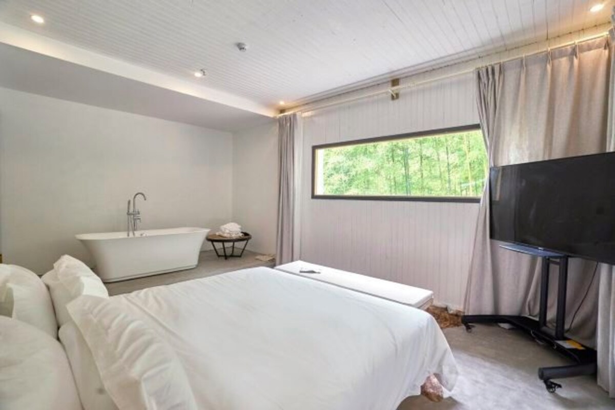 A modern hotel room with a large bed, a bathtub, a television, and a window with a forest view.