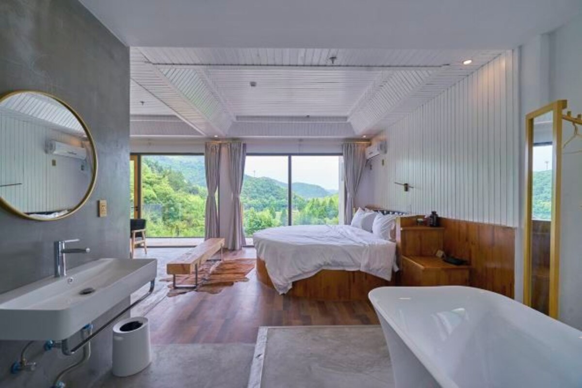 A modern bedroom with a large bed, a bathtub, and a view of greenery.