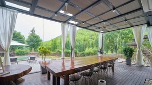 A spacious outdoor dining area with a wooden table and chairs, surrounded by large windows offering a view of a lush garden and mountains. - Rest Valley (Deqing)