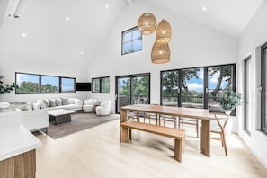 A spacious living area with a large wooden dining table, white sofas, and a view of the outdoors through large windows.