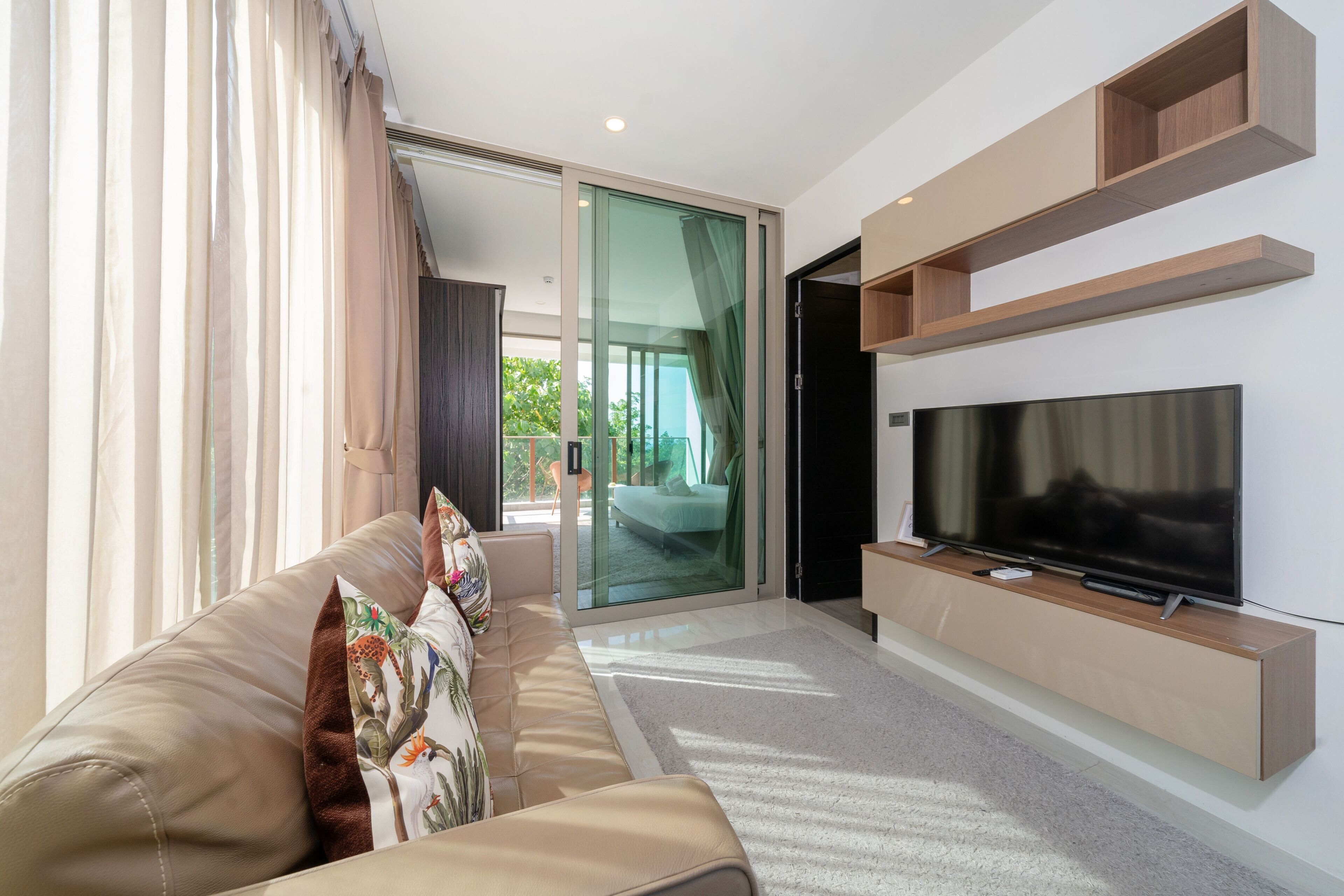 Una sala de estar moderna con un televisor de pantalla plana grande, un sofá color beige con cojines decorativos y un ventanal corredero que da a un balcón.