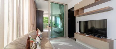 Una sala de estar moderna con un televisor de pantalla plana grande, un sofá color beige con cojines decorativos y un ventanal corredero que da a un balcón.
