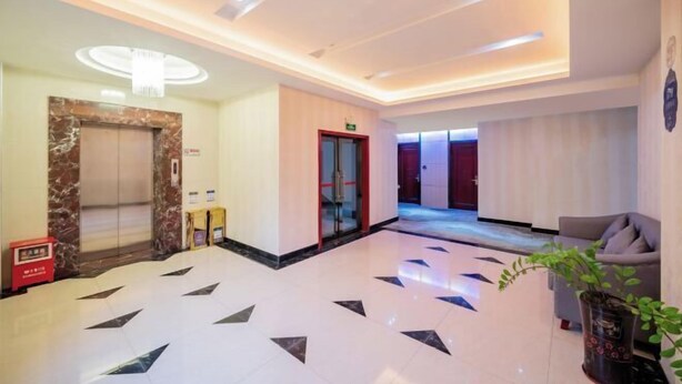 Een hotel lobby met een marmeren receptiebalie, een rode nooduitgangsbord, een glazen lift en een kamerplant.