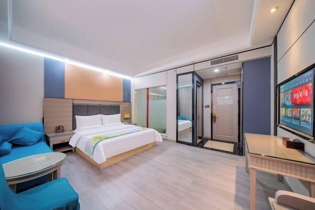Ein modernes Hotelzimmer mit einem Bett, einem blauen Sofa, einem kleinen Tisch, einem Fernseher und einer Schiebetür zu einem weiteren Raum.