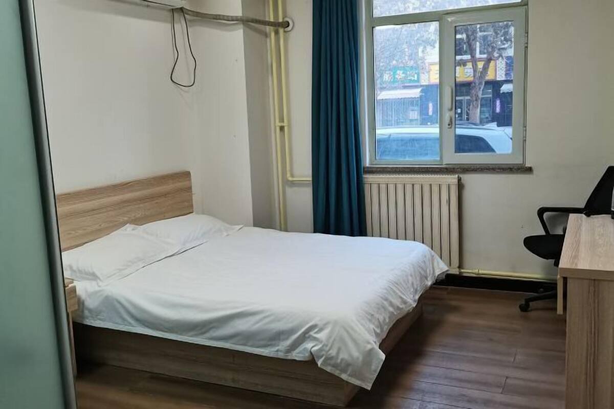 A single bed with a wooden headboard, a window with a view of the street, and a desk with a chair.