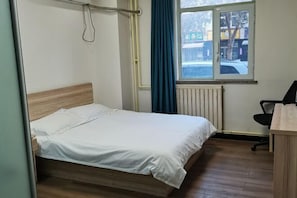 A single bed with a wooden headboard, a window with blue curtains, a radiator, and a desk with a chair. - Jinyuan Express Hotel (Beijing)