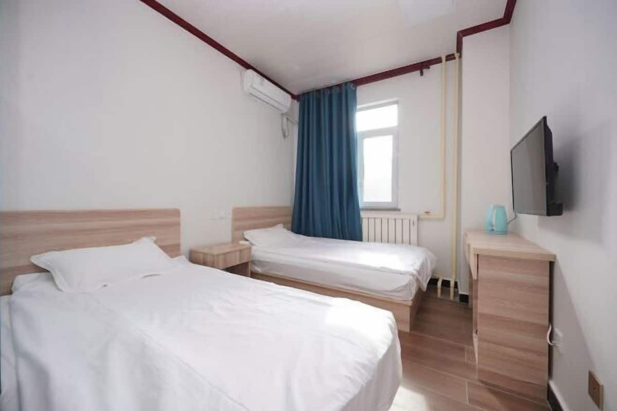A hotel room with two single beds, a wooden headboard, a small bedside table, a TV, and a window with curtains.