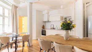 A modern kitchen with white cabinets, a wooden dining table, and chairs. There is a vase with flowers on the table. - Noble - Fantastic 5-Bdr Apt in the Capital (København)