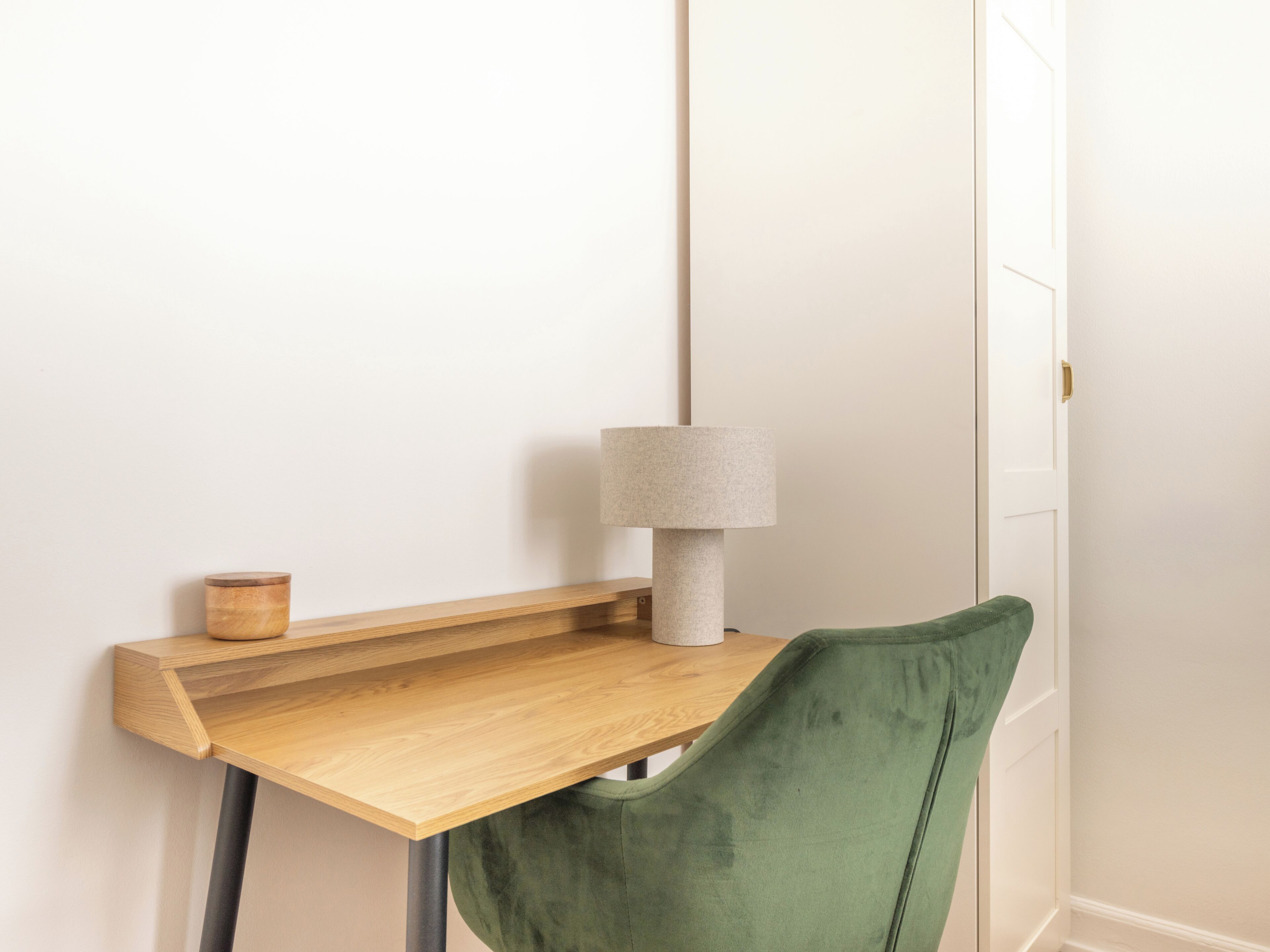 A corner of a room with a wooden desk, a green chair, a lamp, and a small wooden container.