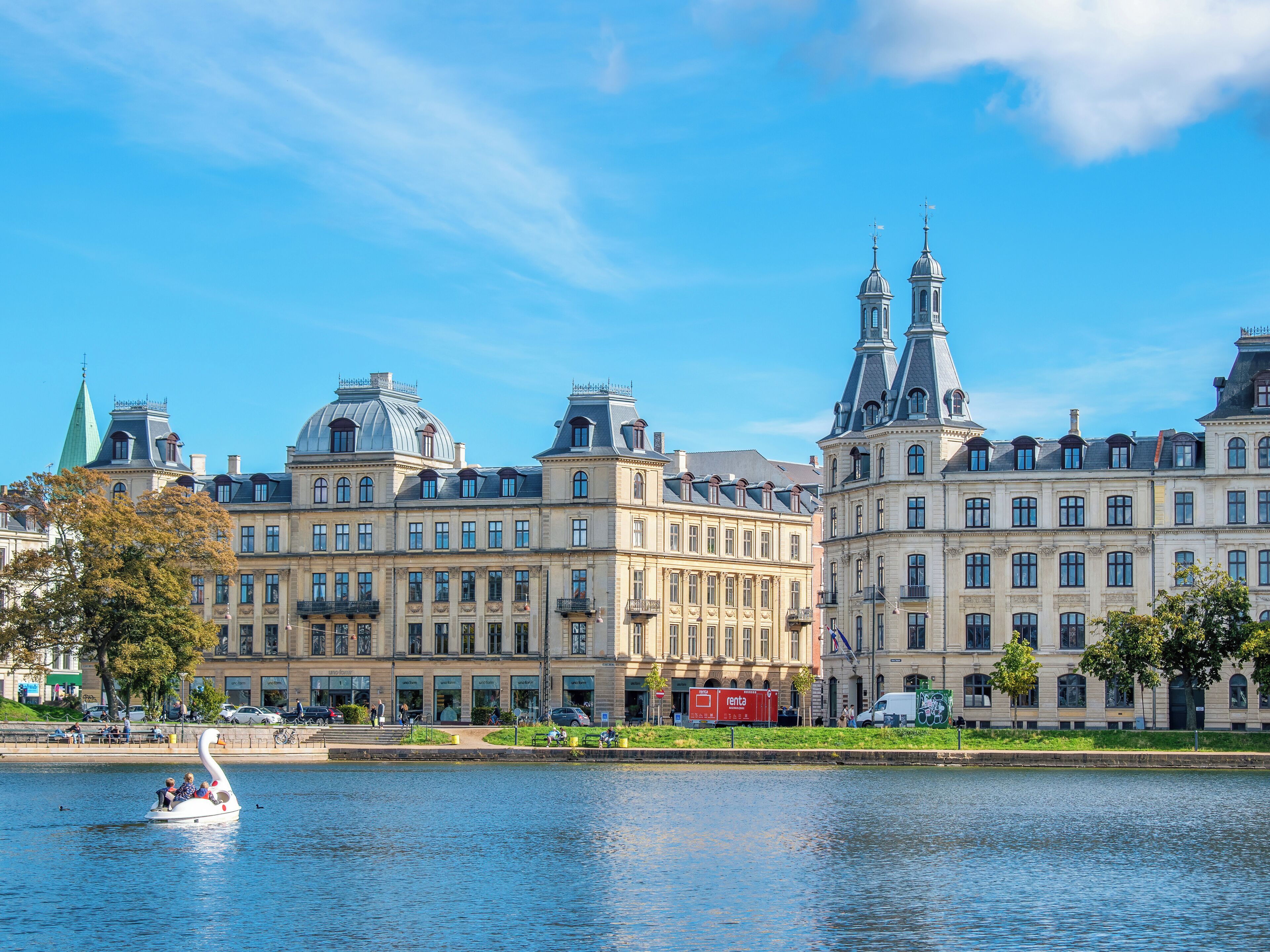 A historic building with classical architecture, a river in the foreground, and a swan-shaped boat with people.